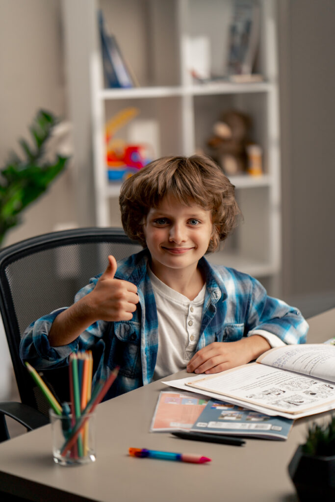 Nahaufnahme im Kinderzimmer: Ein Schüler in weißem Pullover und blau kariertem Hemd sitzt am Schreibtisch, macht Hausaufgaben, schreibt in ein Heft und schaut in die Kamera