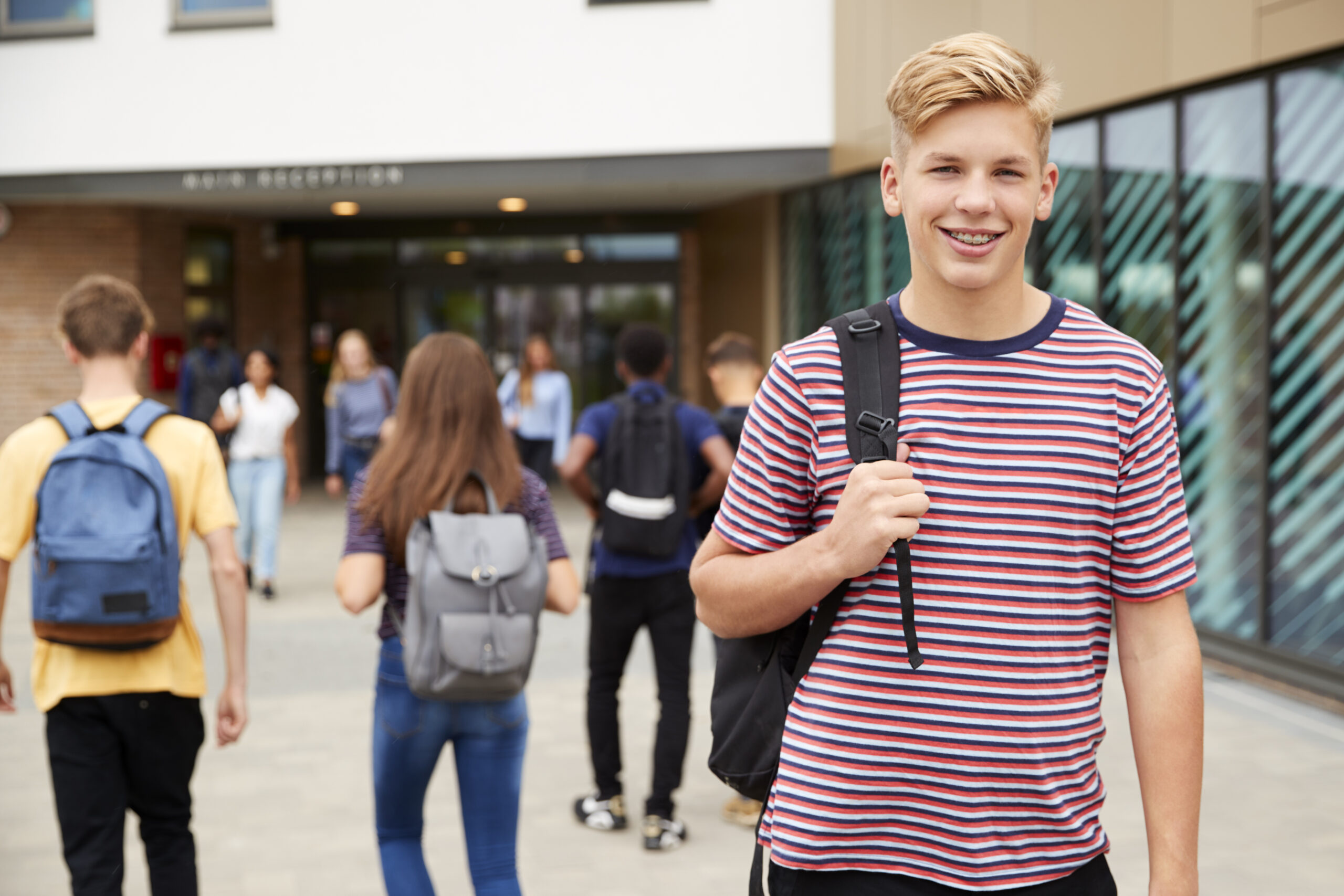 Porträt eines lächelnden männlichen Highschool-Schülers vor einem Schulgebäude, mit anderen Jugendlichen im Hintergrund